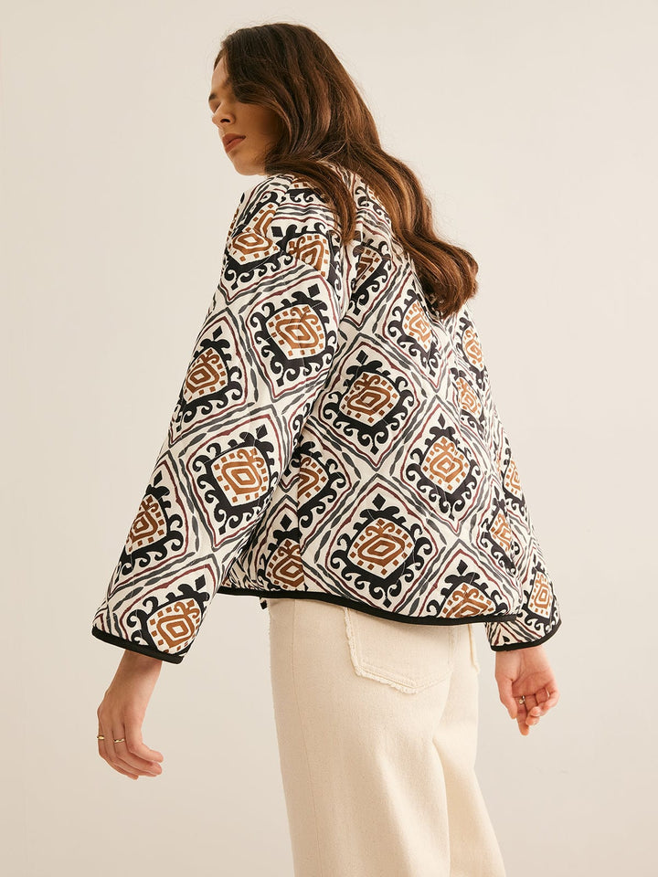 veste ample à motif géométrique noir, blanc et brun, manches bouffantes, bord noir, portée ouverte sur un haut clair et un pantalon clair dans un fond neutre.