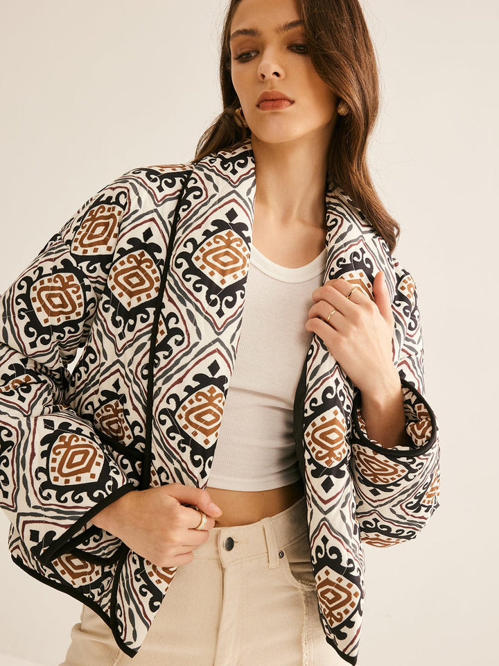 veste ample imprimée de motifs géométriques noir, blanc et brun, manches légèrement bouffantes et bordure noire, portée ouverte sur un haut clair et un pantalon clair dans un cadre neutre.