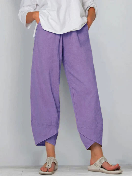 Pantalon droit pour femme de couleur violette, coupe ample et fluide, taille haute avec plis discrets, longueur cheville avec ourlets croisés, porté avec une chemise blanche et des sandales plates, vue de face du buste jusqu’aux pieds sur fond clair.
