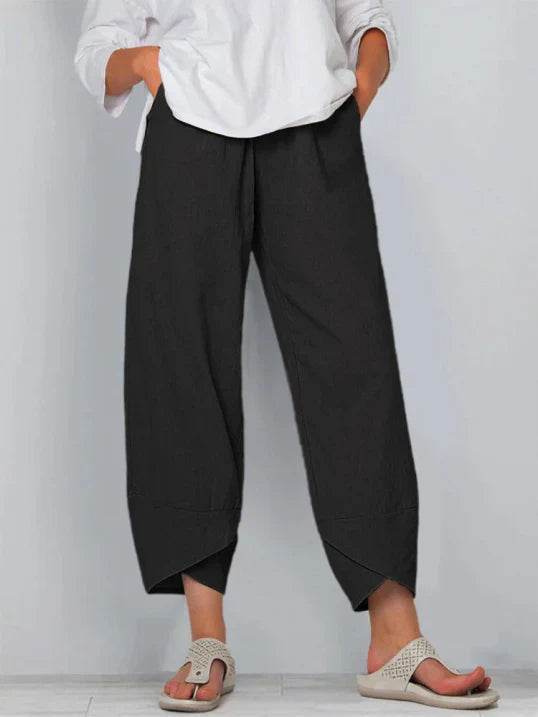 Pantalon droit pour femme de couleur noire, coupe ample et fluide, taille haute avec plis discrets, longueur cheville avec ourlets croisés, porté avec une chemise blanche et des sandales plates, vue de face du buste jusqu’aux pieds sur fond clair.