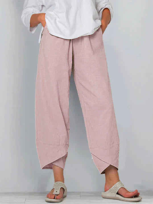 Pantalon droit pour femme de couleur rose poudré, coupe ample et fluide, taille haute avec plis discrets, longueur cheville avec ourlets croisés, porté avec une chemise blanche et des sandales plates, vue de face du buste jusqu’aux pieds sur fond clair.
