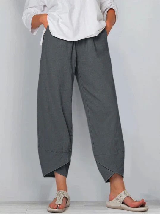 Pantalon droit pour femme de couleur gris foncé, coupe ample et fluide, taille haute avec plis discrets, longueur cheville avec ourlets croisés, porté avec une chemise blanche et des sandales plates, vue de face du buste jusqu’aux pieds sur fond clair.