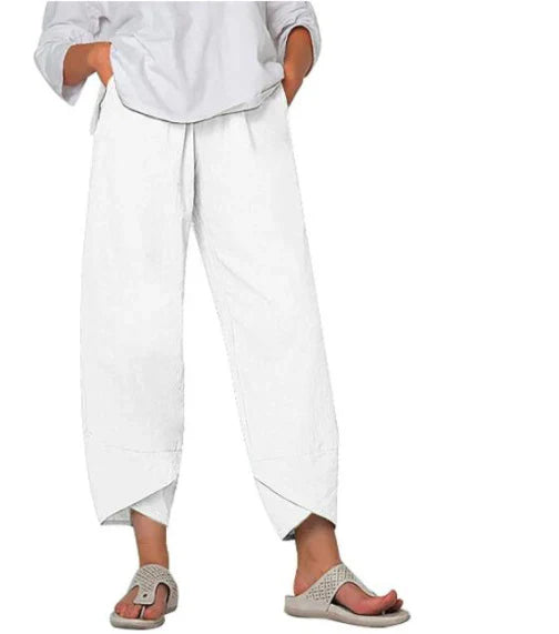 Pantalon droit pour femme de couleur blanche, coupe ample et fluide, taille haute avec plis discrets, longueur cheville avec ourlets croisés, porté avec une chemise claire et des sandales plates, vue de face du buste jusqu’aux pieds sur fond blanc.