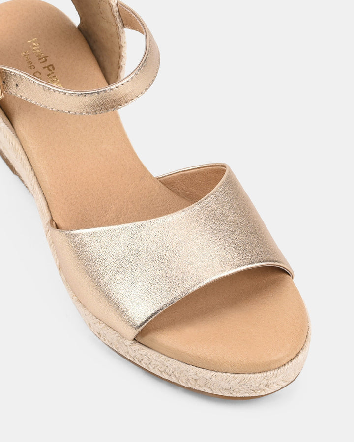 Gros plan sur des sandales compensées dorées pour femmes, mettant en valeur la bride avant large au fini métallisé et la semelle intérieure confortable. Détail de la finition tressée style espadrille pour un look moderne et élégant.