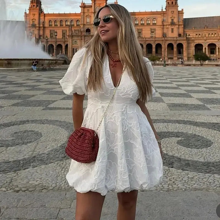 Adoptez un look moderne et raffiné avec la mini-robe Salomé à imprimé floral. Sa coupe féminine avec des manches bouffantes et son décolleté en V en font un choix parfait pour les journées estivales. Ici, elle est portée avec un sac à main en crochet, ajoutant une touche bohème chic à ce style élégant.