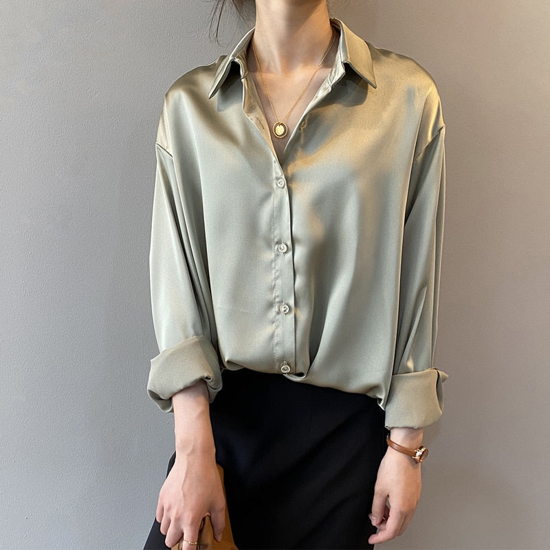 Une femme porte une chemise satinée de couleur vert olive avec un col classique et des manches retroussées. Le haut est boutonné, et elle porte un pantalon noir à taille haute. Elle ajoute une touche élégante avec un collier délicat en or et un sac marron. Son look est raffiné et moderne.