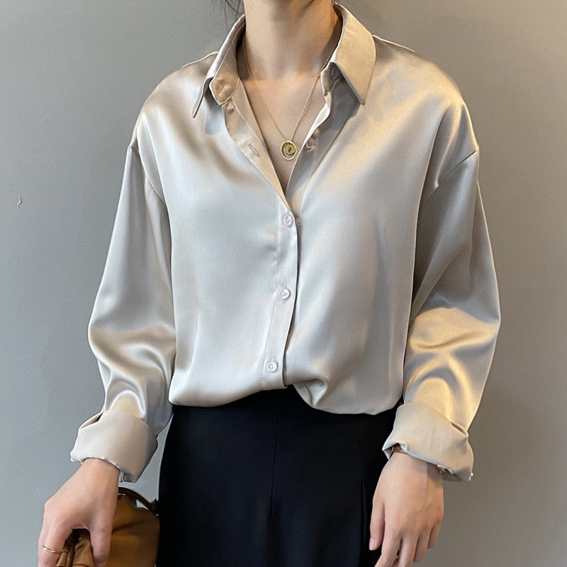 Une femme porte une chemise satinée beige avec un col classique. Les manches sont retroussées et la chemise est légèrement boutonnée, accompagnée d'un pantalon noir à taille haute. Elle porte également un collier délicat en or et tient un sac marron dans ses mains, ajoutant une touche chic à son ensemble.