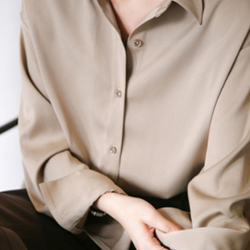 Un gros plan d'une chemise beige en satin avec un col classique. Les boutons sont visibles sur la partie avant, et les manches sont légèrement retroussées. La personne porte également une montre élégante qui complète le look soigné et intemporel.