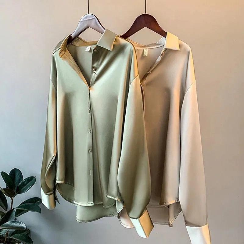 Deux chemises en satin élégantes accrochées sur des cintres. L'une est de couleur vert olive, tandis que l'autre est de couleur beige clair. Les deux chemises présentent un col classique et des manches longues, avec des poignets légèrement retroussés.