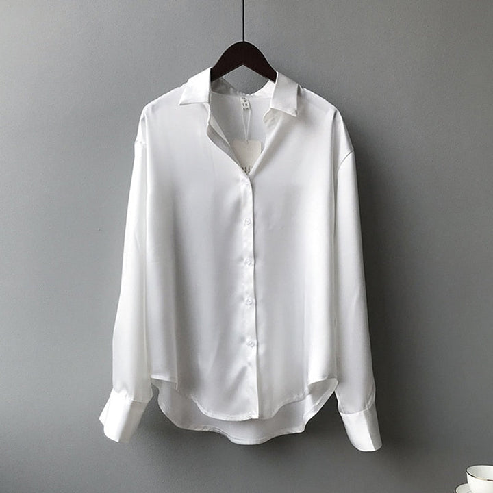 Une chemise blanche en satin élégante accrochée à un cintre. La chemise présente un col classique, des manches longues et des poignets boutonnés. Elle a une coupe fluide et élégante, idéale pour une tenue chic.