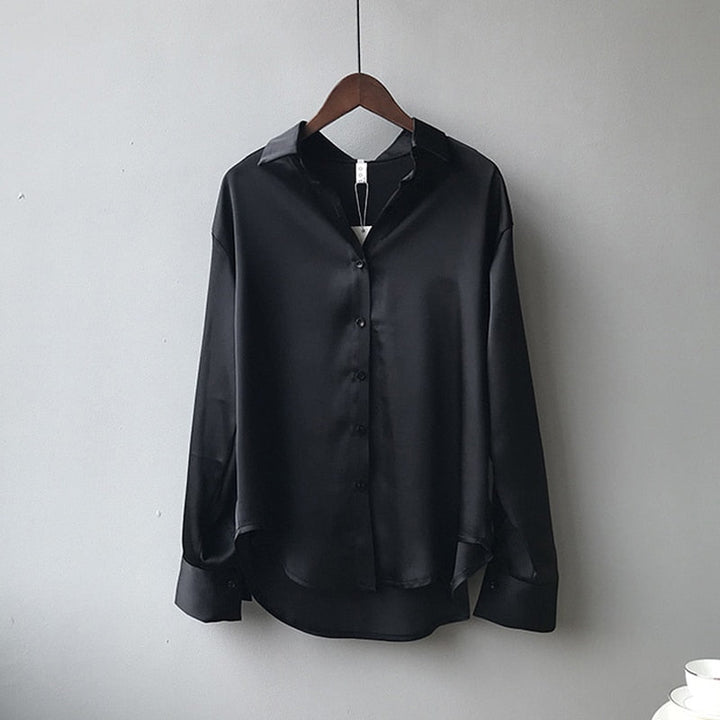 Une chemise en satin noir élégante accrochée à un cintre. Elle possède un col classique, des manches longues avec des poignets boutonnés et une coupe fluide qui lui confère un style moderne et sophistiqué.