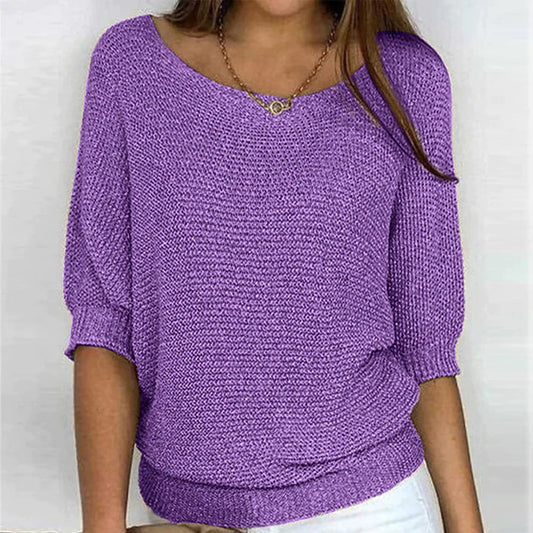 Une femme porte un haut tricoté violet vif à manches 3/4 et un col rond large. Le haut est associé à un pantalon blanc et agrémenté d'un simple collier en or, créant un look à la fois chic et décontracté.
