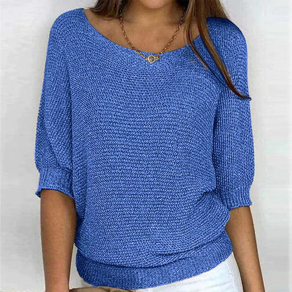 Une femme porte un haut tricoté bleu vif à manches 3/4 et une coupe décontractée. Le haut a un col rond large et est associé à un pantalon blanc. Un collier en or avec un pendentif rond ajoute une touche d'élégance à cette tenue décontractée et chic.