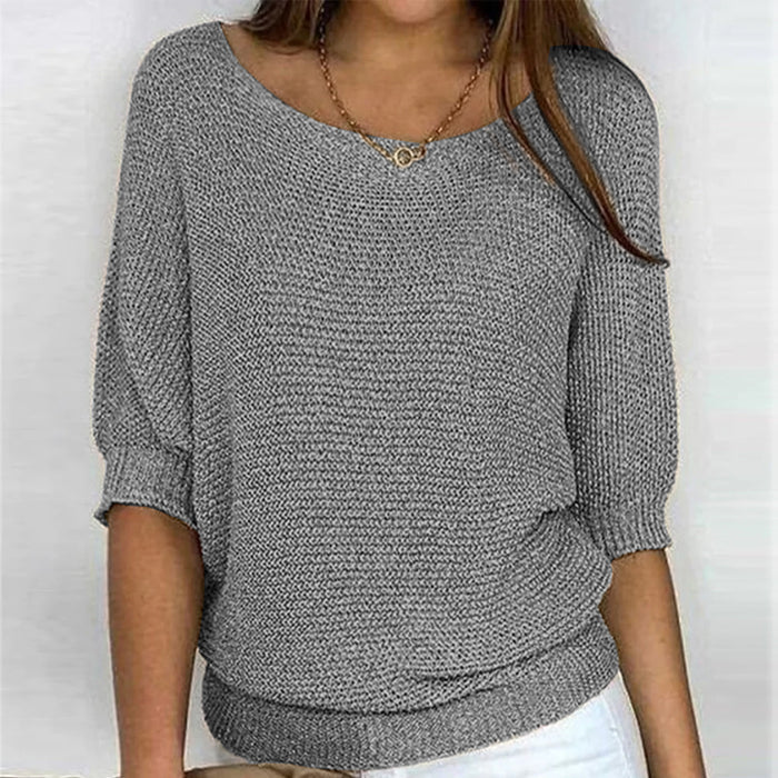 Une femme porte un haut tricoté gris à manches 3/4 et à coupe décontractée. Le haut présente un col rond large, associé à un pantalon blanc et un simple collier en or. Le look est chic et décontracté, offrant une apparence à la fois confortable et stylée.