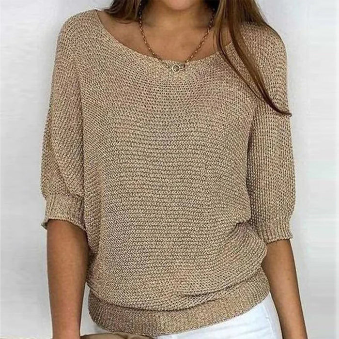 Une femme porte un haut tricoté beige à manches 3/4, avec un col rond large et une coupe décontractée. Le haut est associé à un pantalon blanc et agrémenté d'un collier en or avec un pendentif rond, créant un look à la fois chic et décontracté.