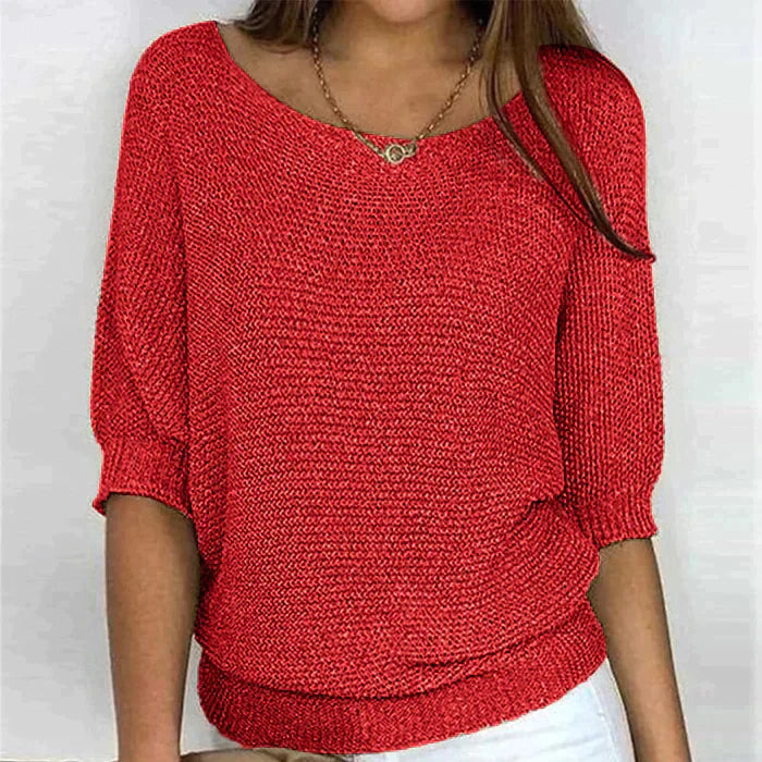 Une femme porte un haut tricoté rouge vif à manches 3/4 et à coupe décontractée. Le haut présente un col en U et un design confortable, associé à un pantalon blanc. Elle accessoirise sa tenue avec un collier en chaîne délicat, ajoutant une touche chic à l'ensemble.