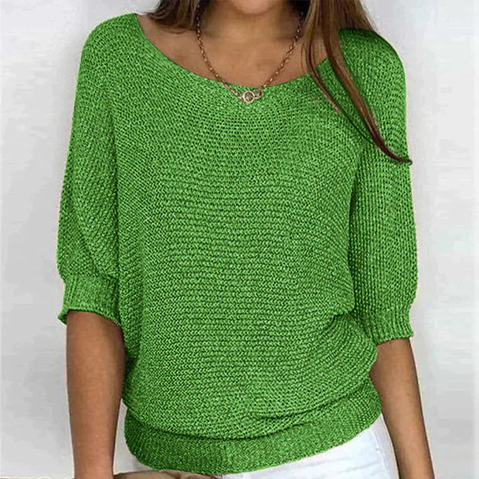 Une femme porte un haut tricoté vert vif à manches 3/4 et à coupe décontractée. Le haut présente un col rond large et est associé à un pantalon blanc. Elle accessoirise sa tenue avec un collier délicat, donnant à l'ensemble un look chic, décontracté et frais.