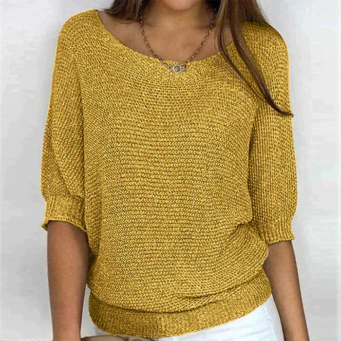 Une femme porte un haut tricoté jaune moutarde élégant à manches 3/4 et à coupe décontractée. Le haut présente un col en U, et elle accessoirise sa tenue avec un collier délicat. Le design chic est associé à un pantalon blanc, créant un look à la fois confortable et tendance.
