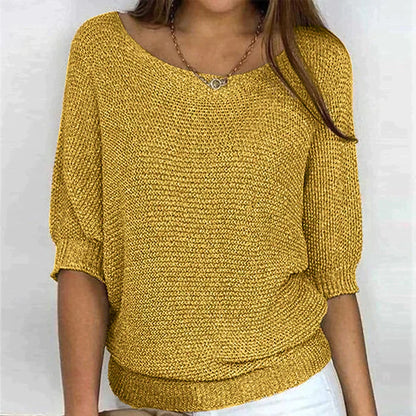 Une femme porte un haut tricoté jaune moutarde élégant à manches 3/4 et à coupe décontractée. Le haut présente un col en U, et elle accessoirise sa tenue avec un collier délicat. Le design chic est associé à un pantalon blanc, créant un look à la fois confortable et tendance.