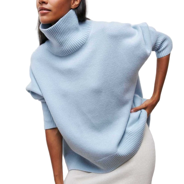 Une femme élégante porte un pull à col roulé ample de couleur bleu pastel. Le vêtement a une texture douce et épaisse avec des bordures côtelées aux manches et à l’ourlet. Elle pose légèrement de profil, une main sur la hanche, mettant en valeur la coupe moderne et décontractée du pull.