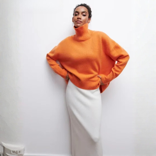 Pull oversize à col roulé pour femme, en orange vif, avec des manches longues et une coupe ample. Le col roulé est large et côtelé, tandis que le bas du pull présente également une finition côtelée. Ce pull est associé à une jupe blanche, créant un look audacieux et moderne, parfait pour une tenue décontractée mais stylée.