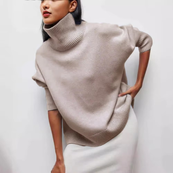 Pull col roulé oversize pour femme, couleur kaki doux, à la coupe ample et aux manches courtes. Le col roulé est large et côtelé, tandis que le bas du pull présente une finition côtelée. Porté avec une jupe blanche, ce pull crée un look à la fois élégant et décontracté, parfait pour une tenue moderne.