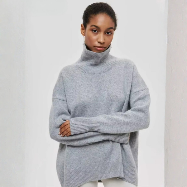 Pull oversize à col roulé pour femme, en gris clair, avec une coupe ample et des manches longues. Le col est haut et côtelé, tandis que le bas du pull présente également une finition côtelée. Le vêtement crée un look à la fois moderne et confortable, idéal pour des tenues décontractées.