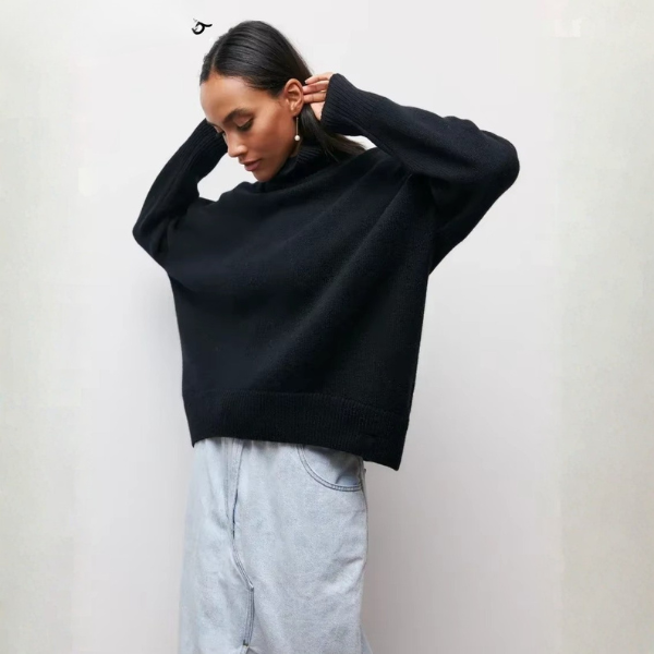 Pull oversize à col roulé pour femme, en noir, avec un ajustement ample et des manches longues. Le pull est porté de manière décontractée, avec une coupe moderne qui tombe au-dessus des hanches, et est associé à une jupe en denim claire, créant un look élégant et confortable.