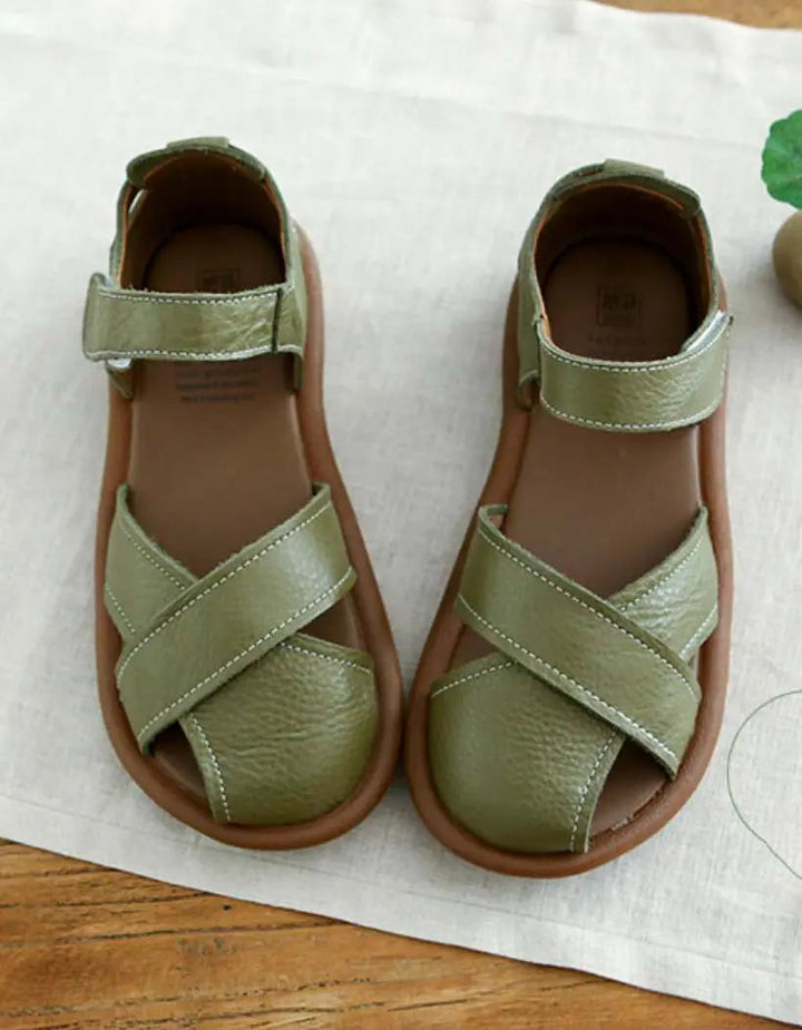 Une paire de sandales plates pour femmes au design moderne, avec des brides croisées en cuir vert olive et une semelle en caoutchouc souple de couleur marron. Ces sandales sont élégantes et confortables, idéales pour les journées estivales.