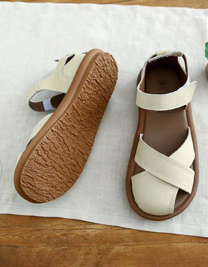 Une paire de sandales plates pour femmes au design moderne, avec des brides croisées en cuir beige clair et une semelle en caoutchouc souple de couleur marron. Ces sandales sont parfaites pour l'été, alliant confort et style.