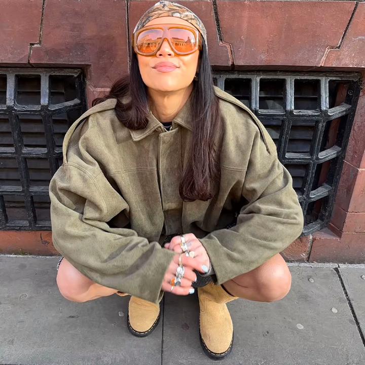 Veste en daim olive au style oversize, portée avec un t-shirt noir, des lunettes de soleil oranges, et des bottines beiges. Un look urbain et décontracté, parfait pour une journée en ville.