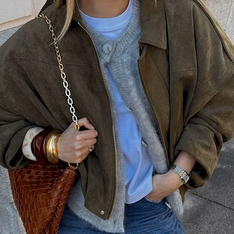 Veste en daim de couleur olive au style décontracté, portée avec un pull gris, un t-shirt blanc et un sac en cuir tressé. Un look chic et confortable pour une tenue quotidienne.