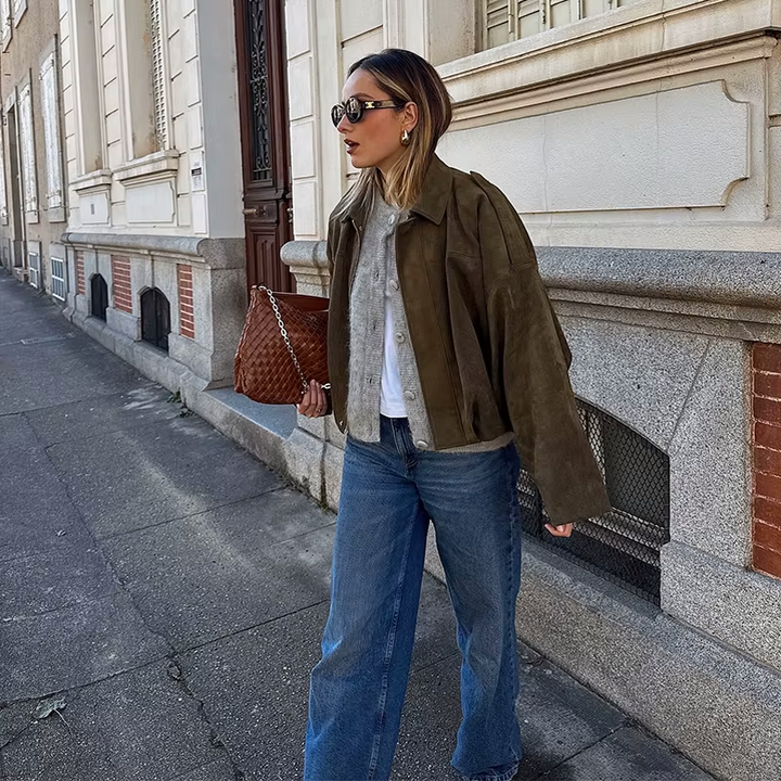 Veste en daim olive portée avec un t-shirt blanc, un cardigan gris, et un jean bleu large. Complétée par un sac à main en cuir tressé et des lunettes de soleil, créant un look chic et décontracté en ville.