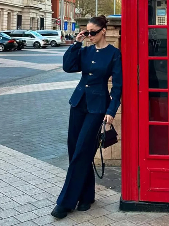 femme en veste en jean bleu foncé à boutons argentés et pantalon assorti, silhouette hivernale chic, en pose urbaine avec sac noir et lunettes de soleil près d’un décor de rue.