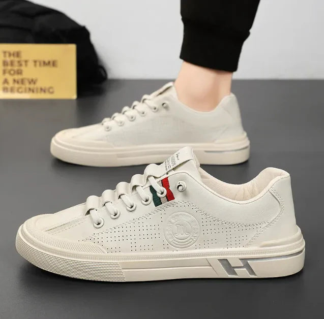Baskets urbaines pour hommes Yvan blanches au style moderne, avec perforations latérales, lacets blancs et détail rouge sur la languette, présentées en studio (une paire portée en arrière-plan.