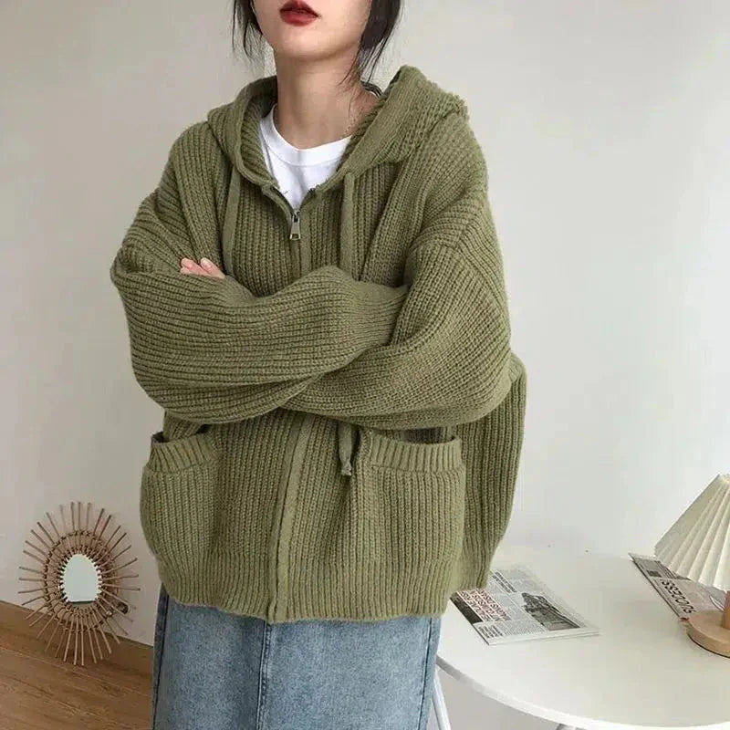 Une femme porte un épais sweat à capuche vert, doux et zippé. Ce vêtement ample possède des poches plaquées sur le devant et des manches longues. Son style décontracté et moderne est parfait pour un look confortable et tendance, idéal pour la mi-saison ou les journées plus fraîches. Le fond minimaliste met en valeur la texture de la maille et la teinte naturelle du sweat.