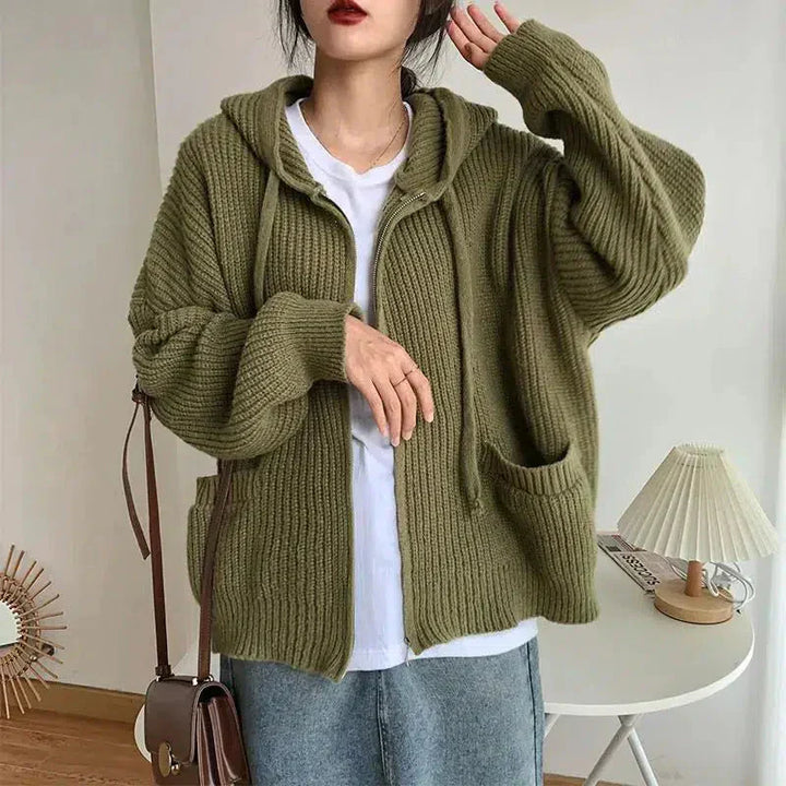 Une femme porte un sweat à capuche vert zippé en maille épaisse et douce. Ce vêtement ample est doté de poches avant et de manches longues légèrement bouffantes. Porté sur un t-shirt blanc et une jupe en jean, il crée un look décontracté et moderne. Son aspect douillet et naturel souligne un style confortable, idéal pour la mi-saison.