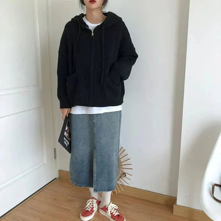 Femme portant un cardigan zippé à capuche de couleur noire, en maille épaisse et douce. Le vêtement présente une coupe oversize avec des poches avant, associé à un t-shirt blanc et une jupe en jean mi-longue. La tenue est complétée par des baskets rouges, créant un look décontracté et moderne. L’ensemble reflète un style confortable et tendance, parfait pour les journées fraîches.