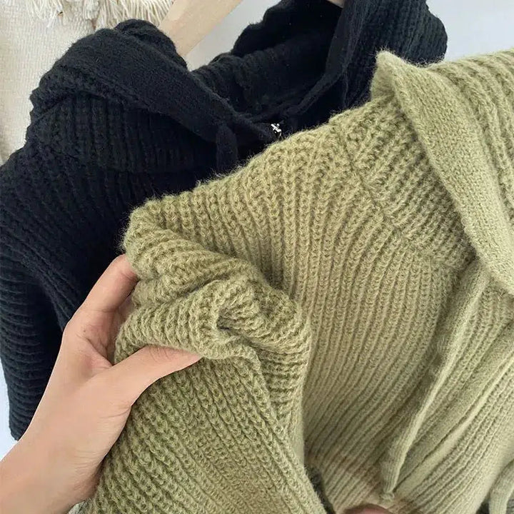 Gros plan sur deux gilets à capuche en grosse maille, l'un vert et l'autre noir. L'image met en valeur la texture douce et moelleuse de la maille, idéale pour un look douillet et chaleureux. Une main tient délicatement le gilet vert, soulignant la qualité et la souplesse du tricot. Les deux pièces dégagent une impression de douceur et d'élégance décontractée.