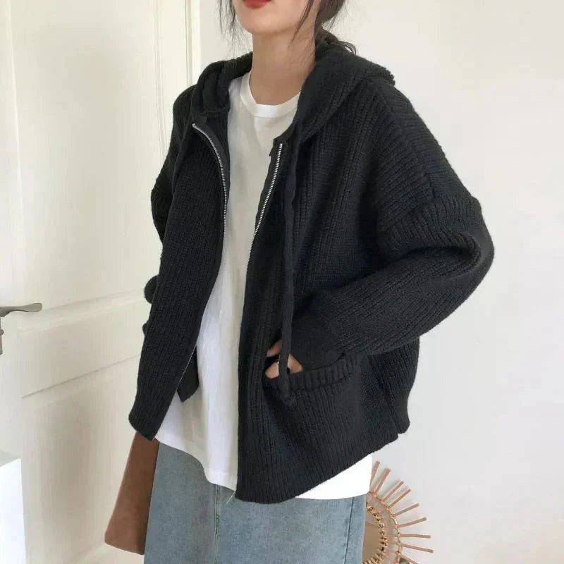 Femme portant un cardigan zippé à capuche de couleur noire, en maille épaisse et douce. Le vêtement présente une coupe oversize avec des poches avant et des manches longues légèrement tombantes. Associé à un t-shirt blanc et une jupe en jean, le look est moderne et décontracté. Le style met en avant un confort optimal et une allure élégante, idéale pour la mi-saison.