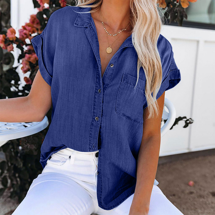 Chemisier en jean bleu foncé pour femme à manches courtes retroussées, coupe moderne et décontractée avec poche poitrine et boutons sur le devant, associé à un pantalon blanc élégant.