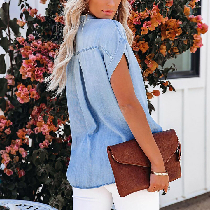 Vue arrière d’un chemisier en jean bleu clair pour femme à manches courtes, coupe moderne et fluide, porté avec un pantalon blanc et une pochette marron pour un style élégant et décontracté.