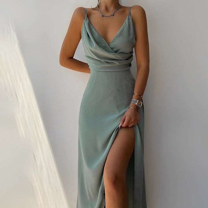 Femme portant une élégante robe longue fluide de couleur vert sauge, à fines bretelles et décolleté drapé. La tenue présente une fente haute sur la jambe, ajoutant une touche sensuelle et sophistiquée. Le tissu satiné reflète délicatement la lumière, créant un effet doux et luxueux, parfait pour une soirée ou un événement chic.