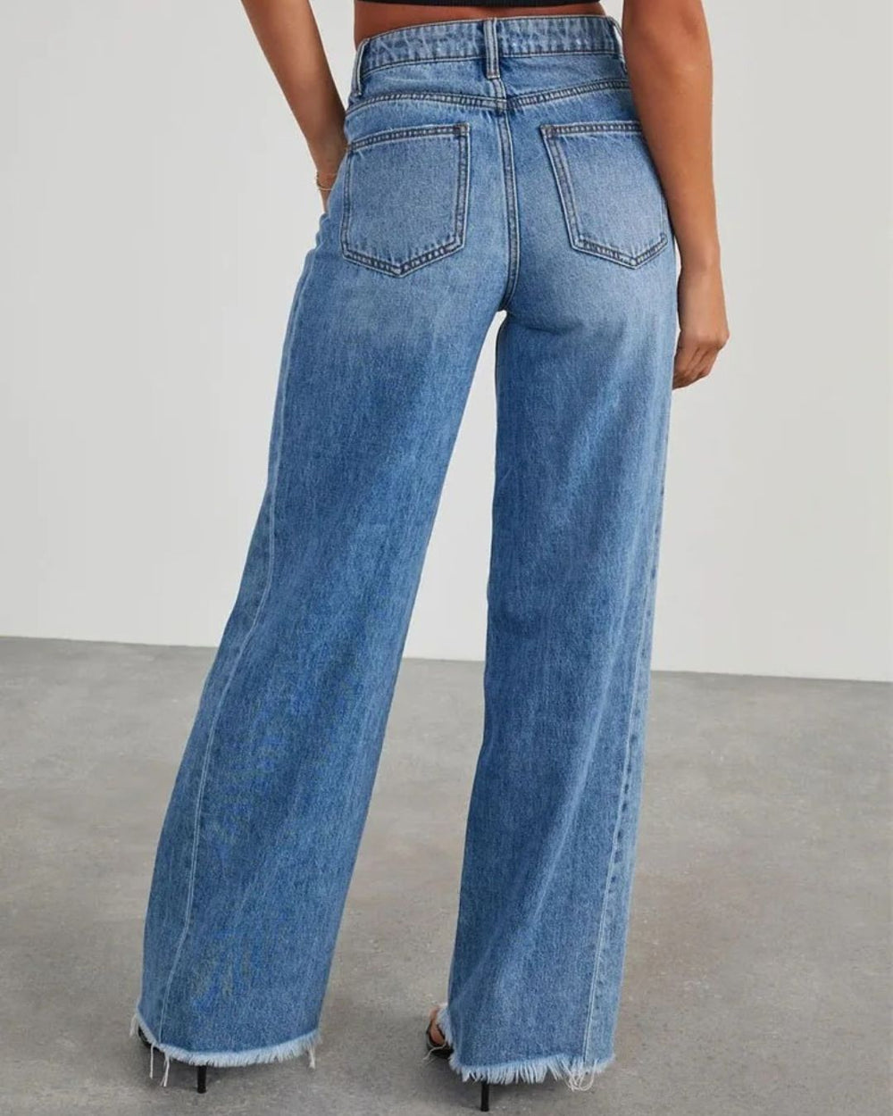Vue de dos du pantalon taille haute Zephyra pour femme en jean bleu, coupe large moderne avec ourlets effilochés, porté avec un haut noir et des sandales à talons.