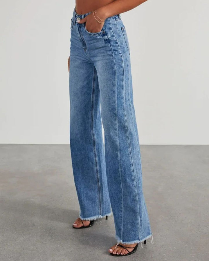 Pantalon taille haute Zephyra pour femme en jean bleu, coupe large moderne avec ourlets effilochés, porté avec des sandales à talons, vue de profil.