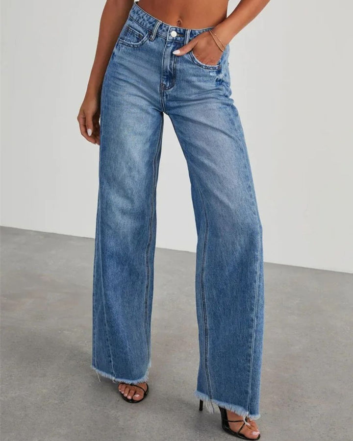Pantalon taille haute Zephyra pour femme en jean bleu délavé, coupe large moderne avec ourlets effilochés, porté avec des sandales à talons, vue de face.