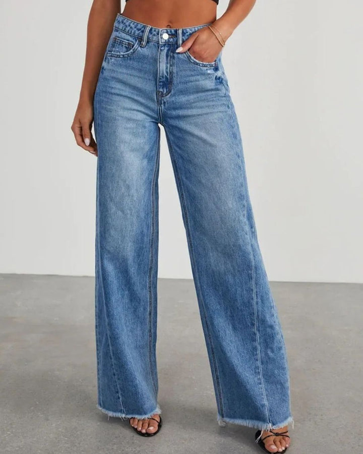 Pantalon taille haute Zephyra pour femme en jean bleu, coupe large moderne avec ourlets effilochés, porté avec un haut court et des sandales à talons, vue de face.