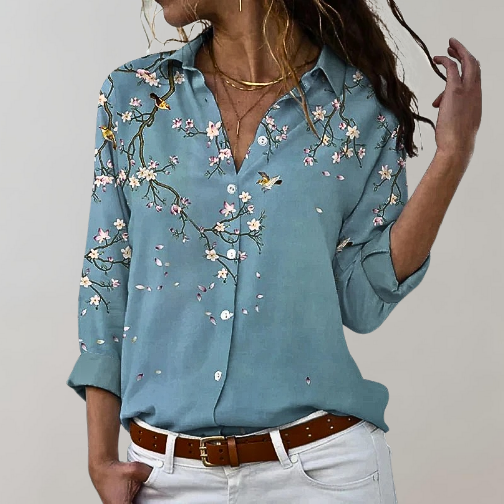 Chemise bleu clair avec motifs floraux et oiseaux, col classique et manches retroussées, élégante et féminine pour un look raffiné.