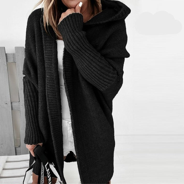 Femme portant un long cardigan noir en maille épaisse, doté d’une capuche et de manches larges, superposé à une tenue blanche, dans un décor lumineux et minimaliste.