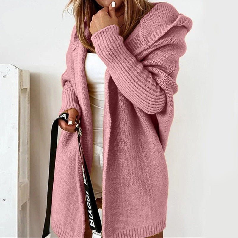 Femme vêtue d’un long cardigan rose en maille douce, avec capuche et manches côtelées, porté par-dessus une tenue blanche dans un décor clair et minimaliste.